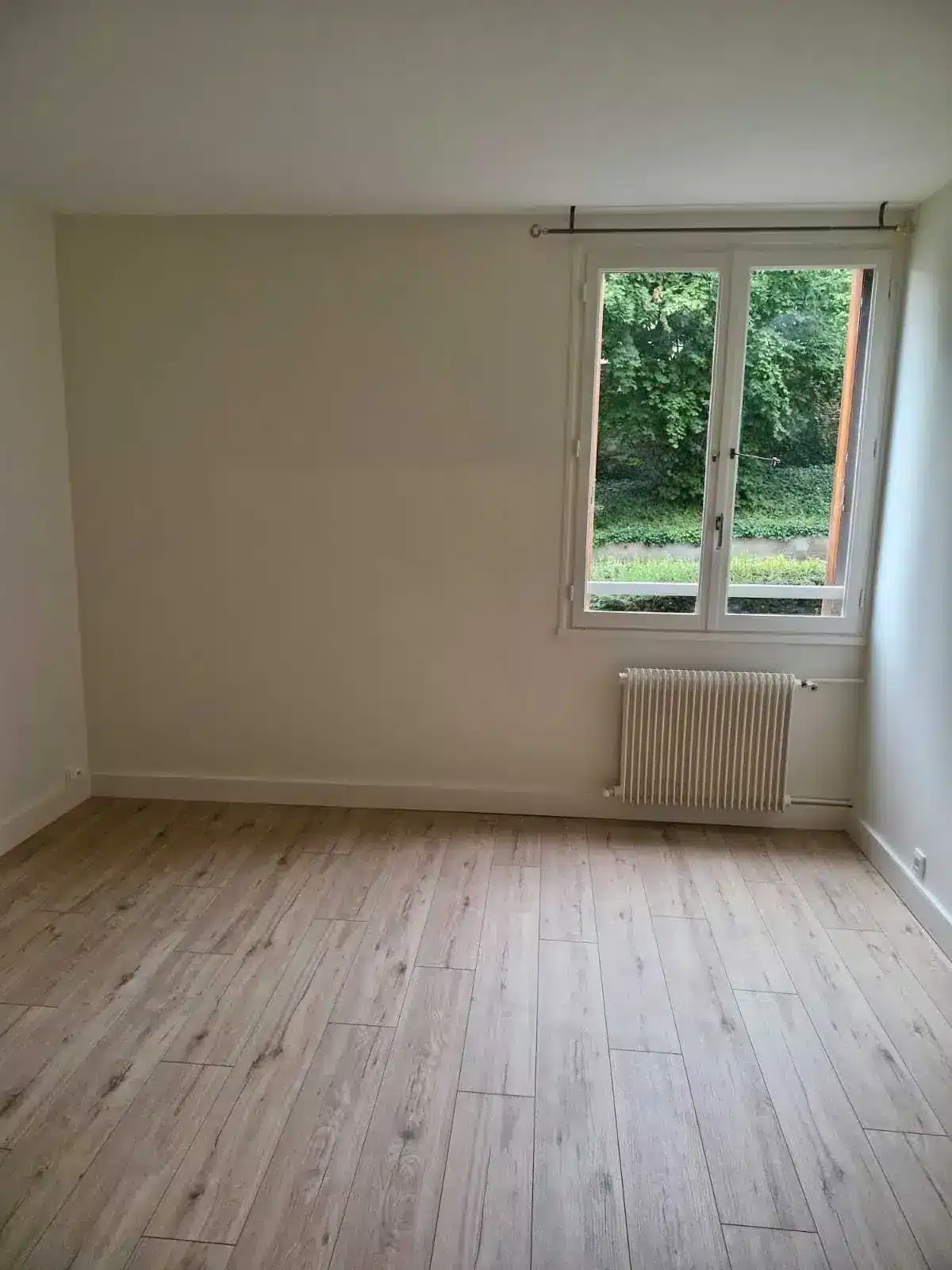 Chambre vide rénovée avec murs blancs, parquet clair, radiateur et fenêtre sur jardin