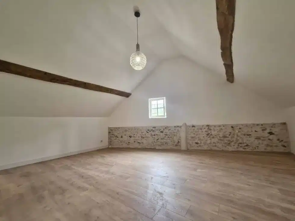 Pièce sous combles avec poutres apparentes, mur en pierre, parquet et murs blancs