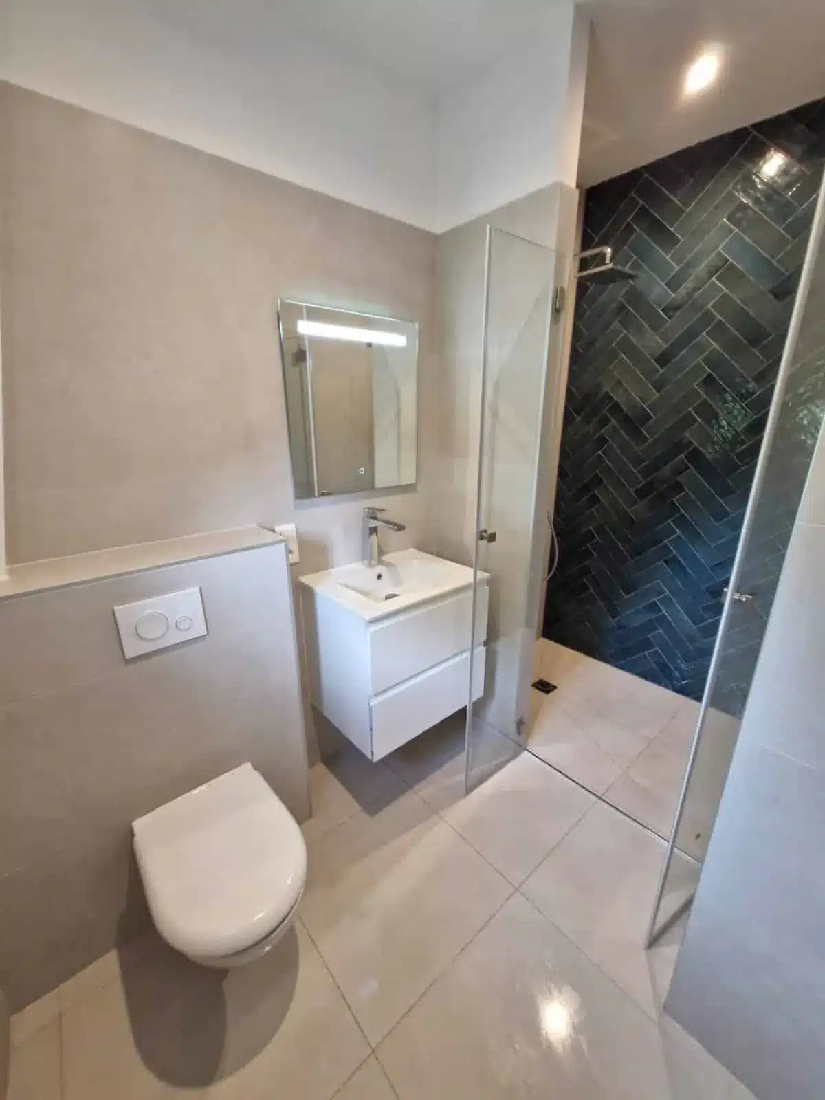 Salle de bain avec meuble vasque blanc, miroir lumineux, WC suspendu et douche en carrelage chevron