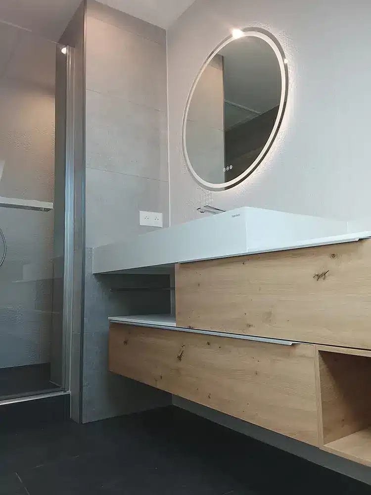 Meuble vasque suspendu en bois clair et miroir rond à éclairage intégré dans une salle d'eau rénovée